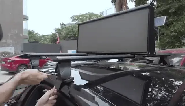 A rooftop LED screen fitted across the crossbars of a dark car parked on a street. An installer's hands are on the clamp at the front bar. Other parked cars and buildings are behind.